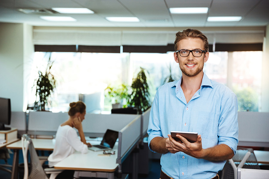 Young handsome successful businessman smiling, holding tablet, office background.